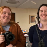 Two girls smile with each other and the girl on the left holds a Canon camera.